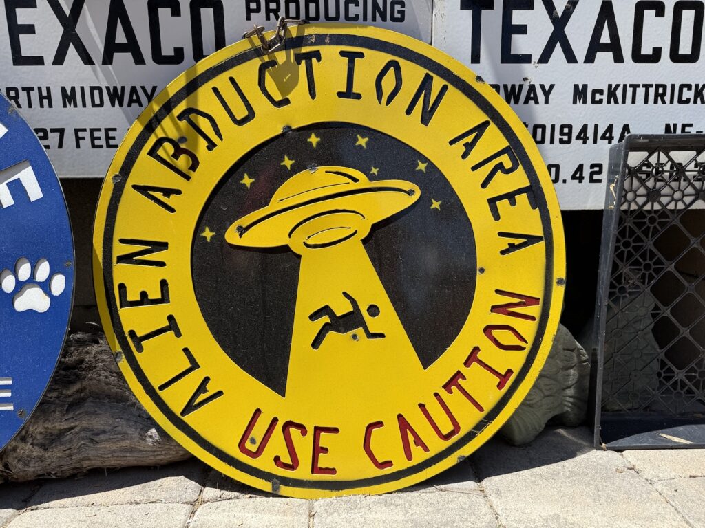 Round, yellow metal sign used for decoration in gardens or walls. The image is of a man being beamed up into a space ship. The text says Alien Abduction Area, use caution
